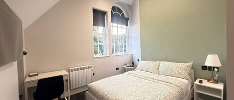 A bedroom with a bed, a desk, a chair, a radiator, a window, and a nightstand with a lamp.