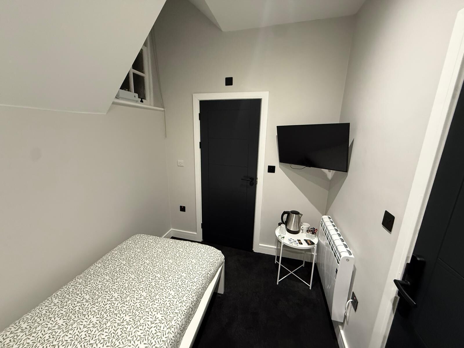 A small, compact room with a single bed, a small table with a kettle and cups, a radiator, and a TV mounted on the wall.