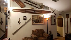 Un soggiorno con soffitto alto, travi in legno, un quadro di una grande barca a vela, un orologio, una lampada e una poltrona.