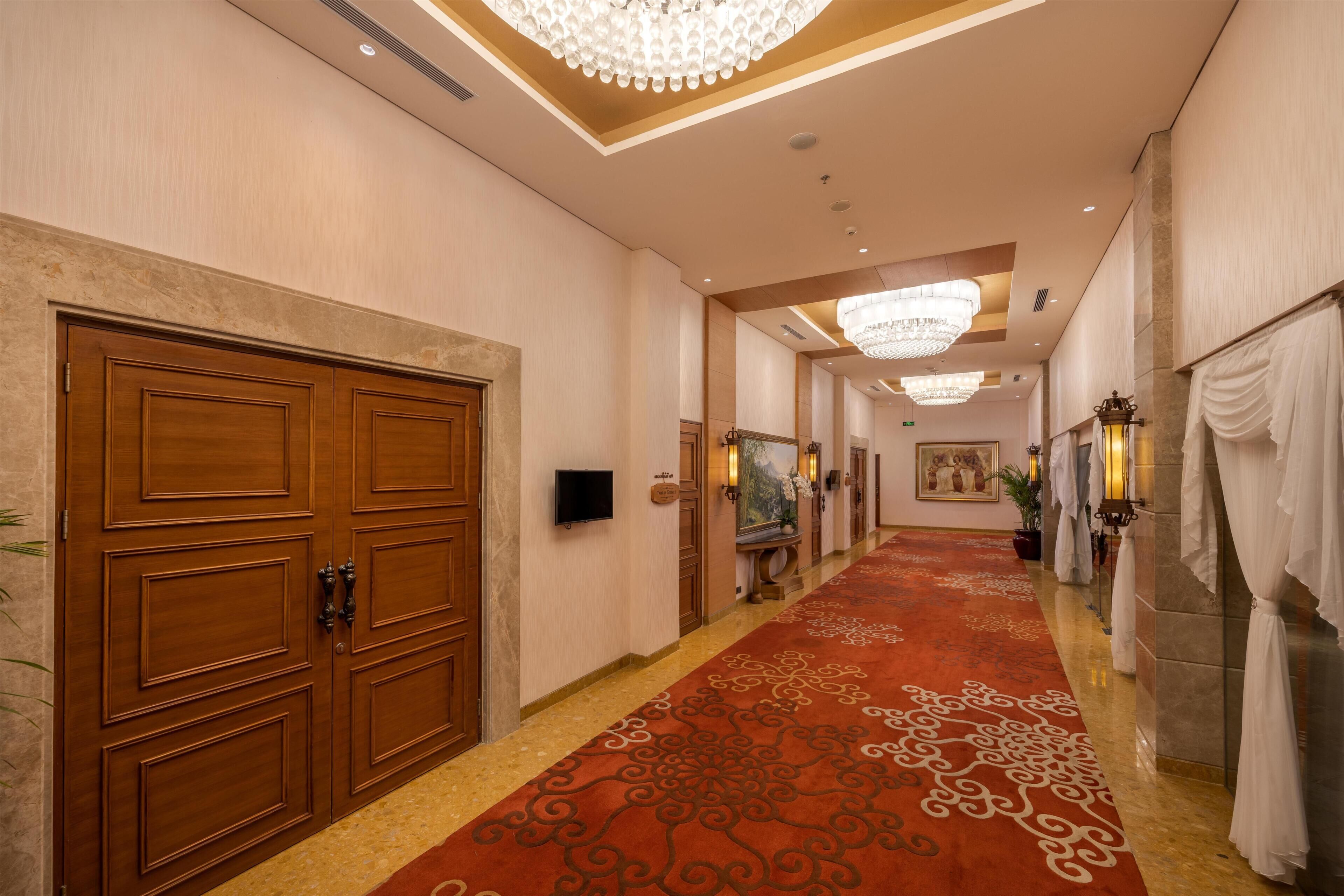 Un pasillo de hotel con puertas de madera, alfombra roja, candelabros y un televisor montado en la pared.