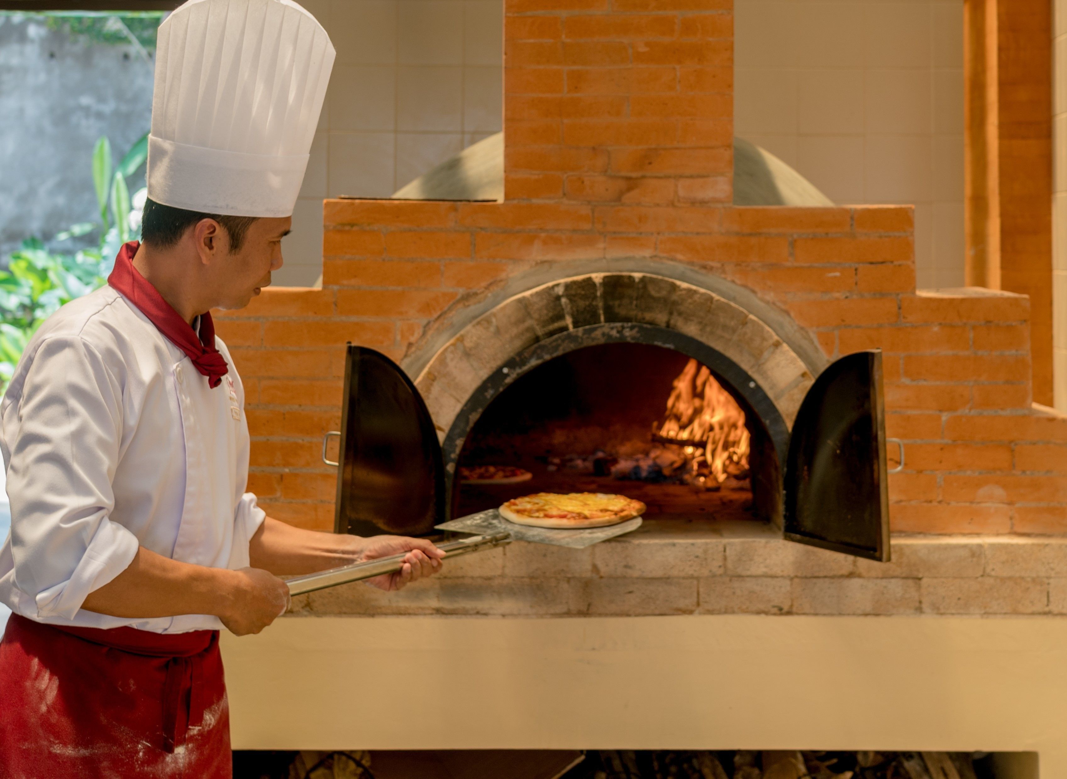 A chef in a white uniform with a red apron, holding a pizza in front of a brick oven.