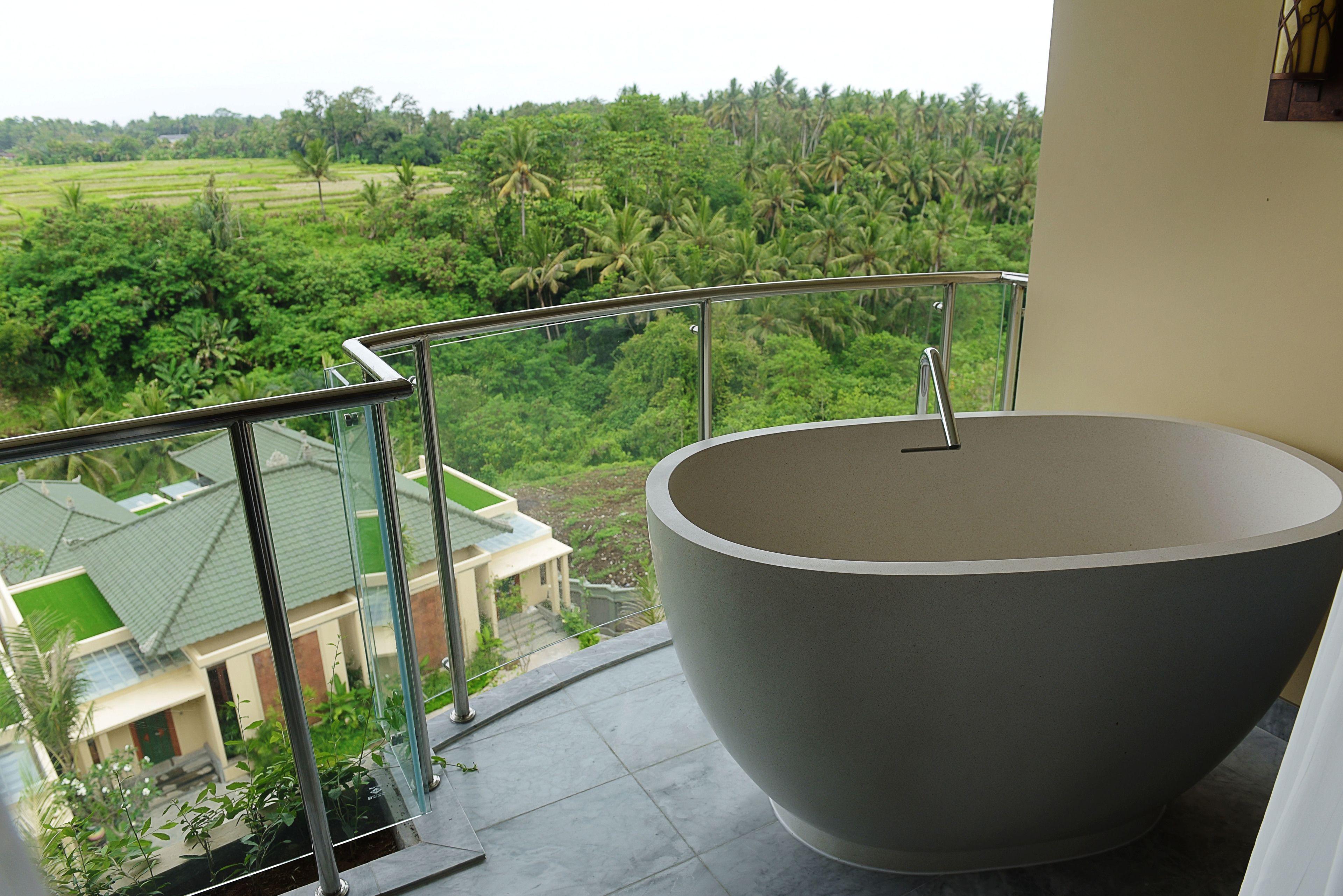 A balcony with a large, modern bathtub and a view of a lush green landscape with palm trees and a house in the background.