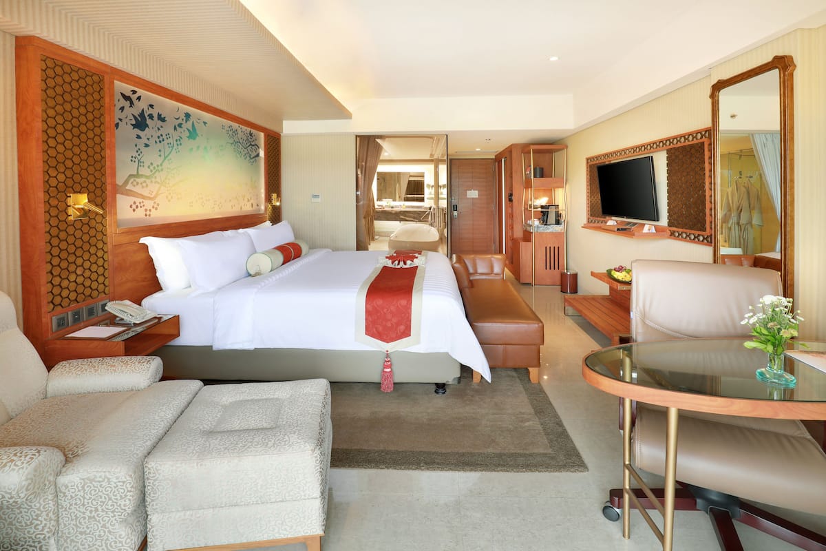 A hotel room with a large bed, a sitting area with a glass table, a TV, and a bathroom visible through an open door.