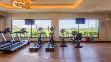 A gym with treadmills, ellipticals, and stationary bikes, large windows with a view of greenery, and ceiling lights.