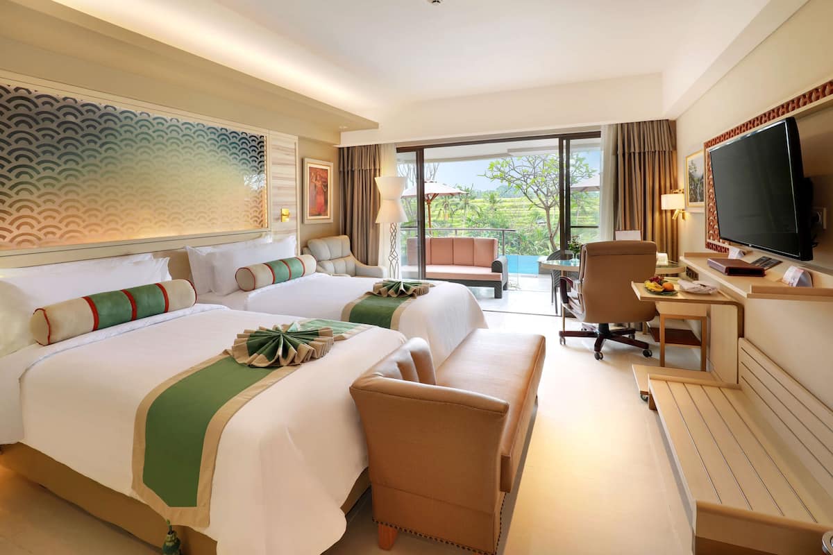 A hotel room with two beds, a desk with a computer, a TV, and a balcony with a view of greenery.