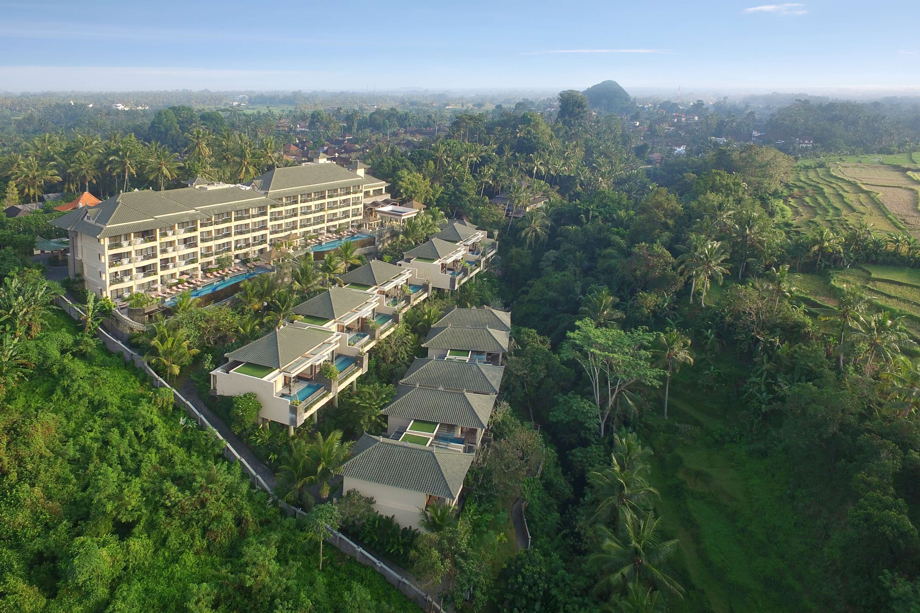 Une vue aérienne d’un complexe hôtelier comprenant plusieurs bâtiments, dont un grand hôtel avec une piscine et des villas plus petites, le tout entouré d’une végétation luxuriante et de palmiers.
