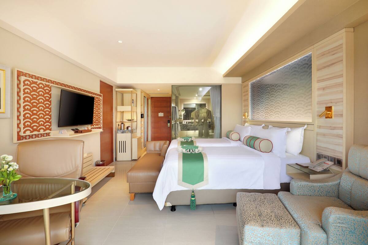 A hotel room with a bed, a TV, a seating area, and a bathroom visible through a glass door.