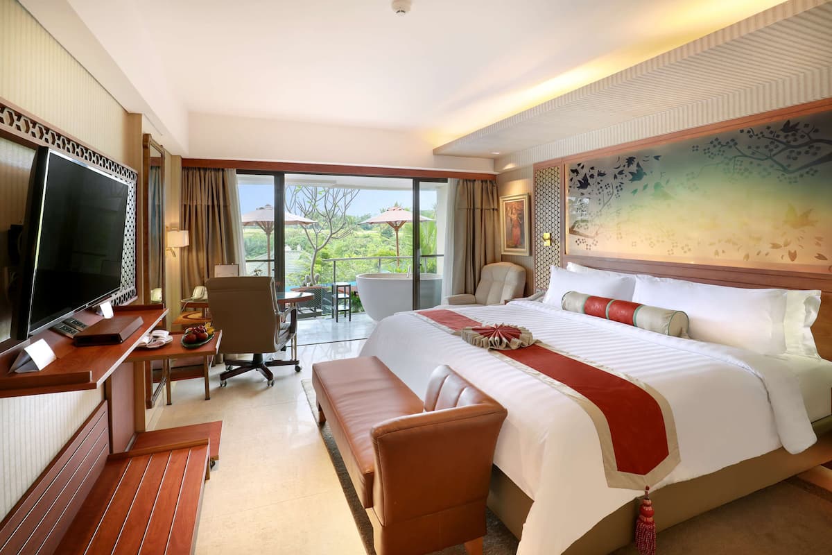 A hotel room with a large bed, a desk with a flat-screen TV, a seating area, and a balcony with a view of greenery.