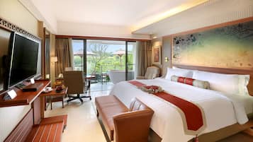 A hotel room with a large bed, a desk with a flat-screen TV, a seating area, and a balcony with a view of greenery.