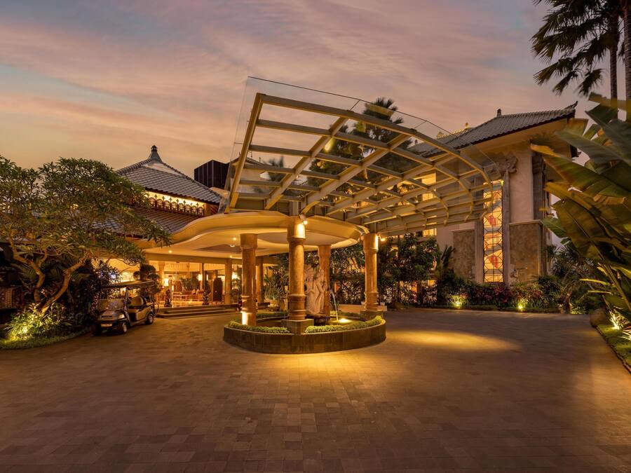 A hotel entrance with a covered walkway, palm trees, and a traditional building in the background.