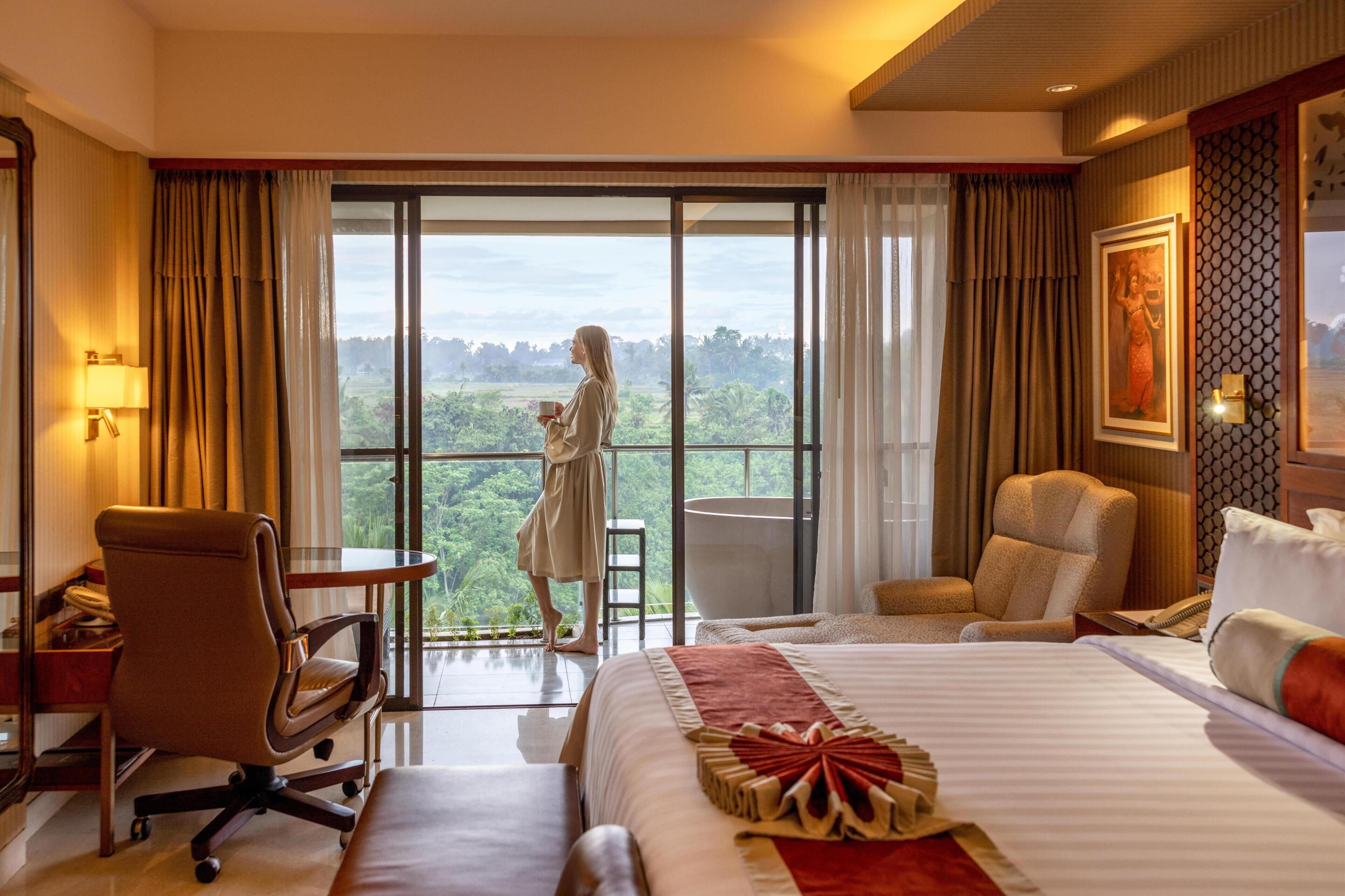 A hotel room with a large bed, a desk, a chair, a sofa, and a view of a lush landscape through a sliding glass door.