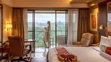 A hotel room with a large bed, a desk, a chair, a sofa, and a view of a lush landscape through a sliding glass door.