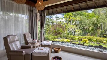 A room with two leather chairs, a small table, and a view of a lush garden through large windows.