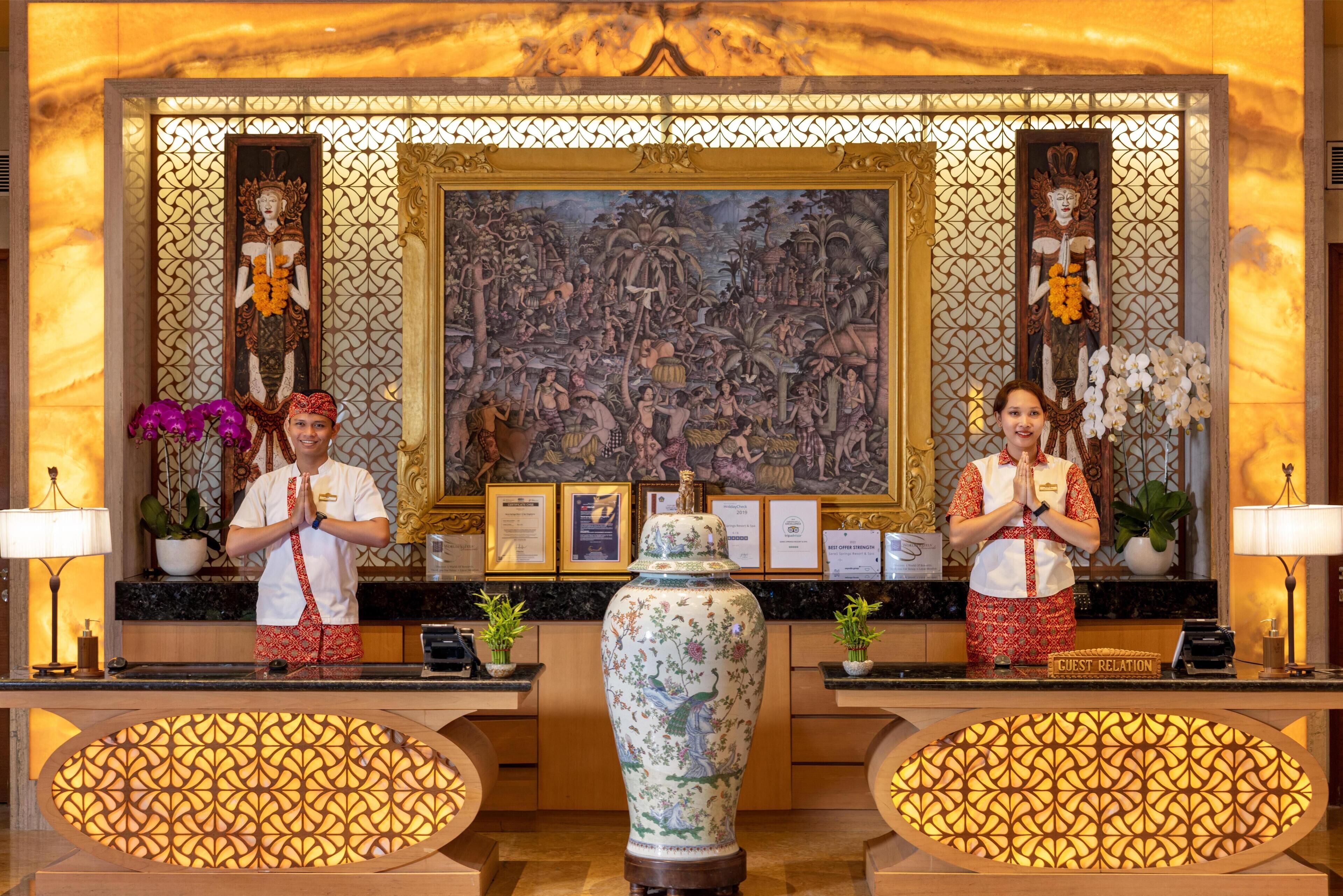Uma área de recepção com um vaso grande, obras de arte emolduradas e uma caixa de atendimento ao hóspede.