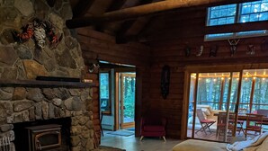 A cozy cabin interior with a stone fireplace, wooden furniture, and a view of the forest through large windows.