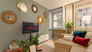 Een woonkamer met een beige bank, een flatscreen televisie, decoratieve spiegels en potplanten.