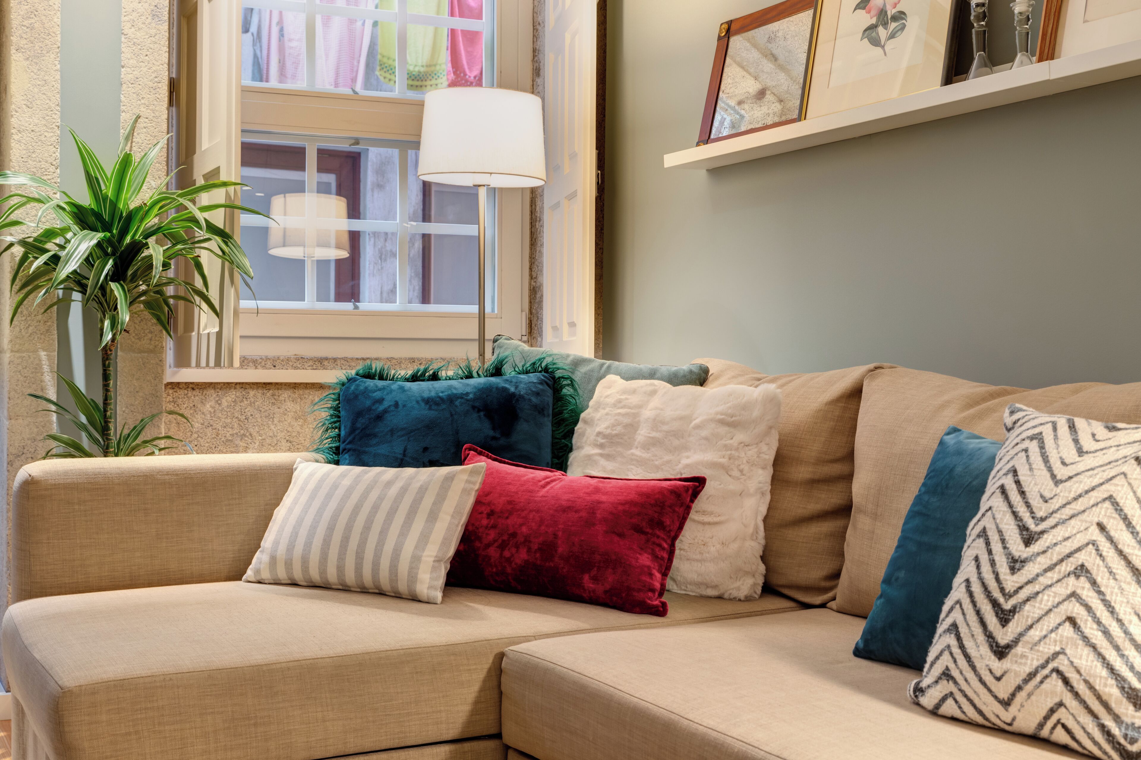 A beige sectional sofa with multiple pillows, a potted plant, and a window with a lamp.
