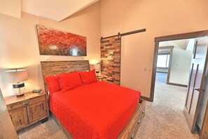 A bedroom with a red bedspread, wooden headboard, a painting on the wall, and a nightstand with a lamp.