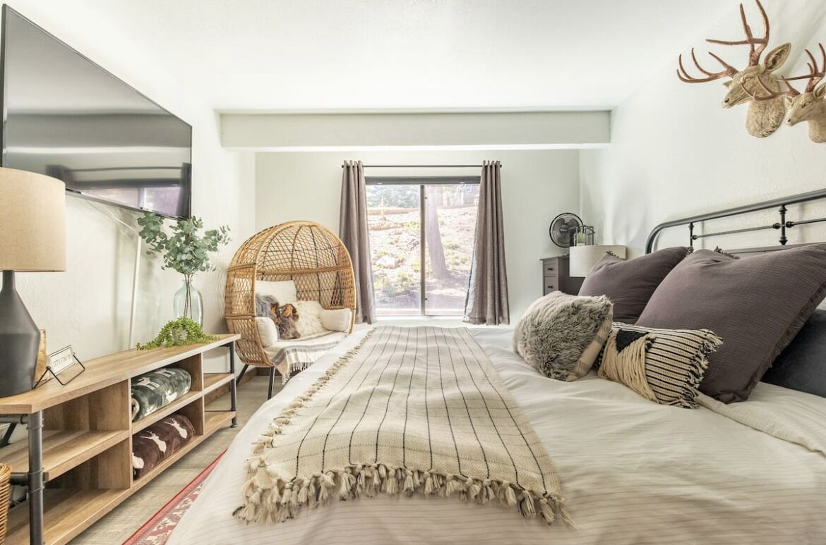 A bedroom with a large bed, a bedside table, a lamp, a wicker chair, a view of the outdoors, and a wall-mounted deer head decoration.