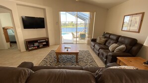 A living room with a brown leather sofa, a wooden coffee table, a flat-screen TV, and a sliding glass door leading to a balcony with a pool. - EAGLE POINTE SOUTH ON THE LARGER LAKE  A NEW VACATION/HOLIDAY EXPERIENCE (Kissimmee)