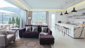 Una sala de estar moderna con sofá, reposapiés y sillones, una cocina con taburetes de bar y ventanas amplias con vistas a árboles.