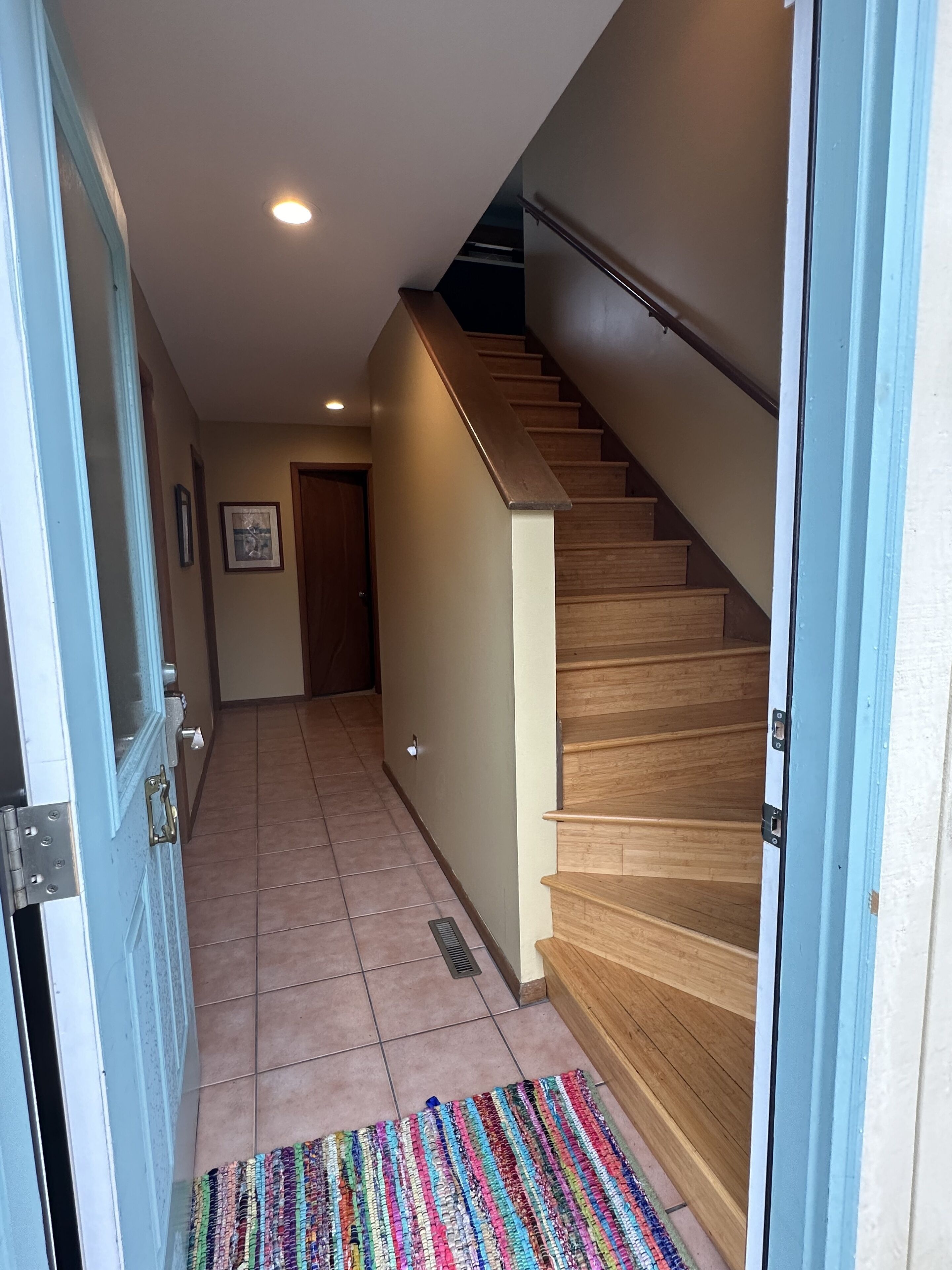 A hallway with a wooden staircase, a tiled floor, and a colorful rug.