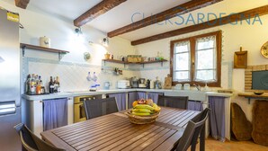 A kitchen with a wooden table, chairs, and a fruit basket. There are shelves above the counter with various items. - Spectacular Villa with independent pool for 8 people near the beach (Alghero Italy)