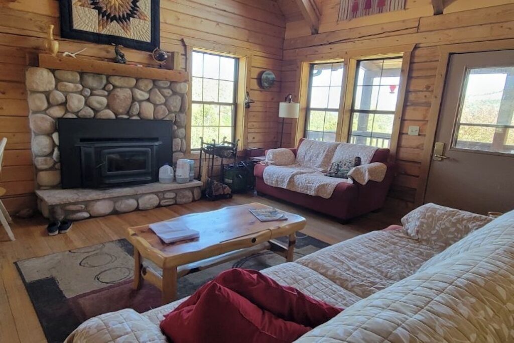 Un interior acogedor de cabaña con chimenea de piedra, un sofá, una mesita de centro y un ventanal grande.