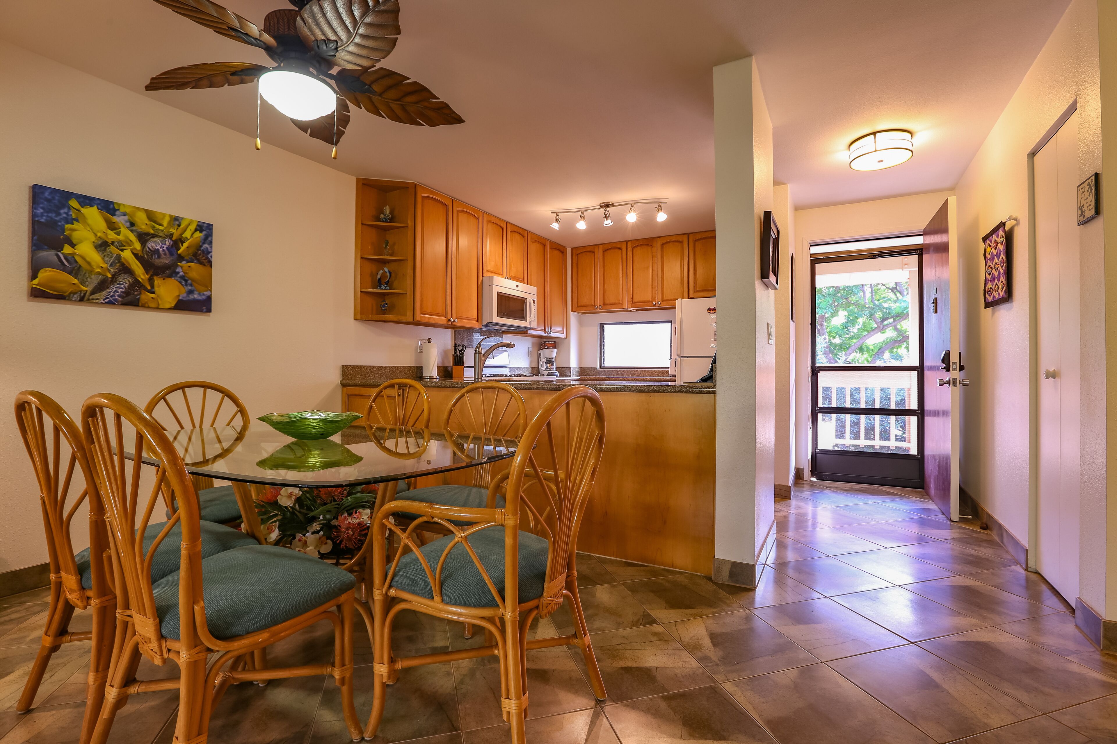 Una cocina con armarios de madera, un microondas y una zona de comedor con sillas de mimbre y una mesa de vidrio.