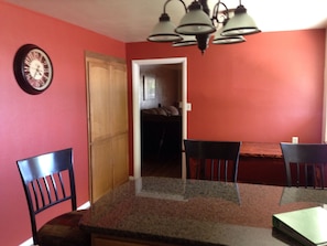 A dining area with a glass table, two chairs, a clock, and a door leading to a bedroom.