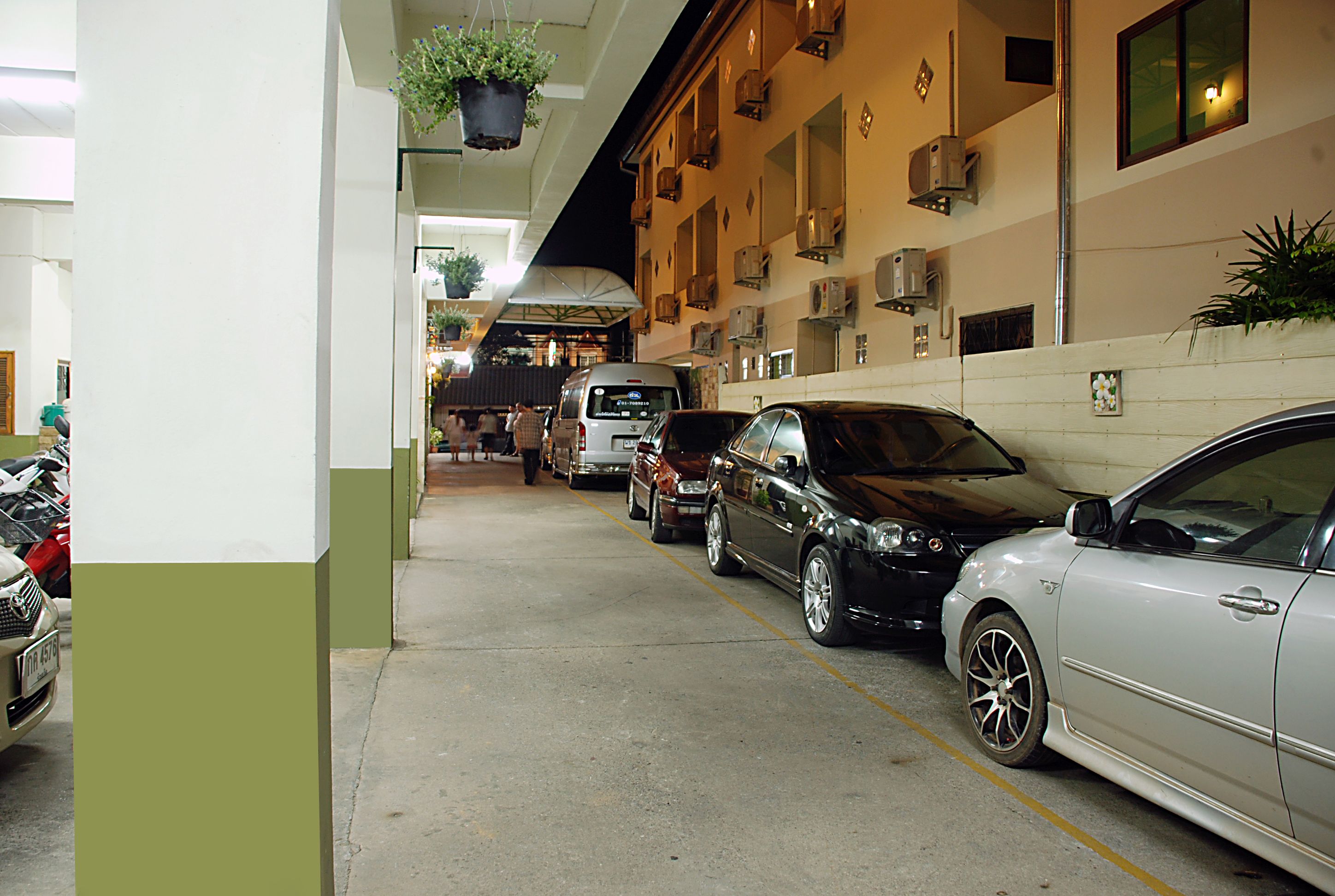 A parking area with multiple cars, a building with air conditioning units, and hanging plants.
