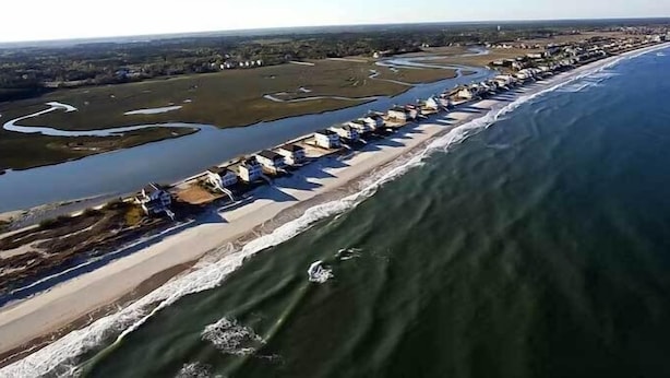 Vista aérea de una zona costera con una larga hilera de casas de playa a lo largo de la orilla, un río o bahía junto a la playa, y un cielo despejado.