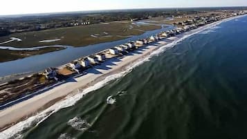 An aerial view of a coastal area with a long row of beach houses along the shoreline, a river or bay adjacent to the beach, and a clear sky.