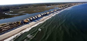 Une vue aérienne d’une zone côtière, avec une longue rangée de maisons de plage le long du rivage, une rivière ou une baie adjacente à la plage, et un ciel dégagé.