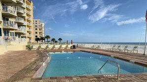 Una piscina con solárium empedrado, vista al océano y un edificio con balcones.