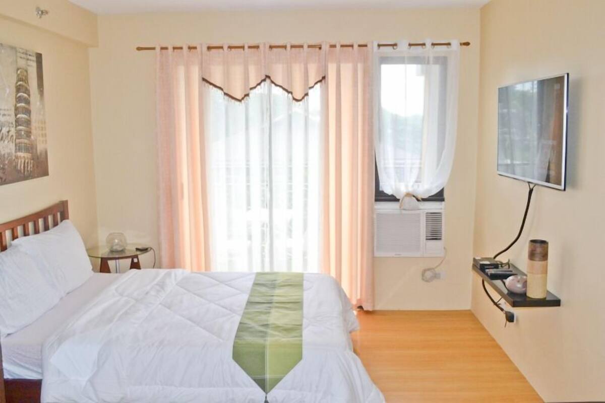 Une chambre d’hôtel avec un lit, un téléviseur fixé au mur, une petite table avec une lampe et une fenêtre avec des rideaux.