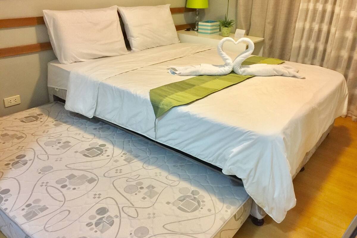 Un lit bien fait, avec du linge de lit blanc et un plaid vert. On peut voir sur le lit une décoration en forme de cœur, réalisée avec des serviettes.