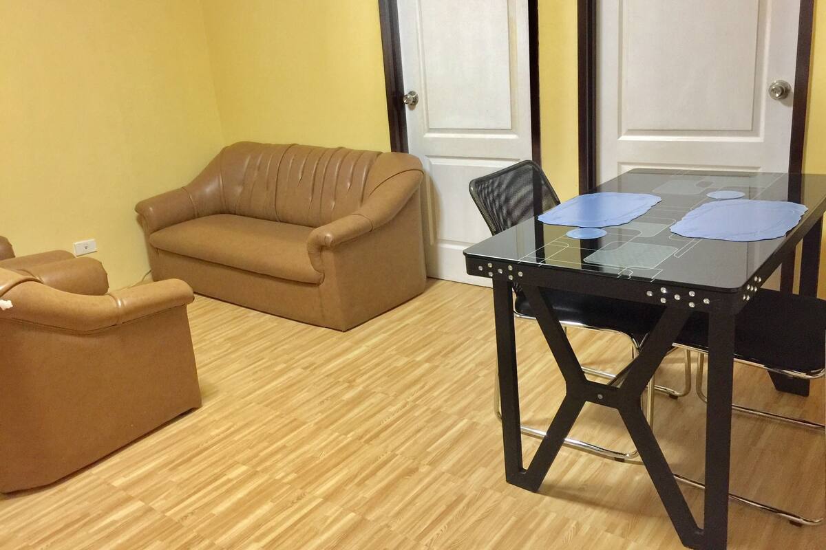 Une pièce avec un canapé en cuir marron, une table à manger en verre et aux pieds en métal, et un plancher en bois.