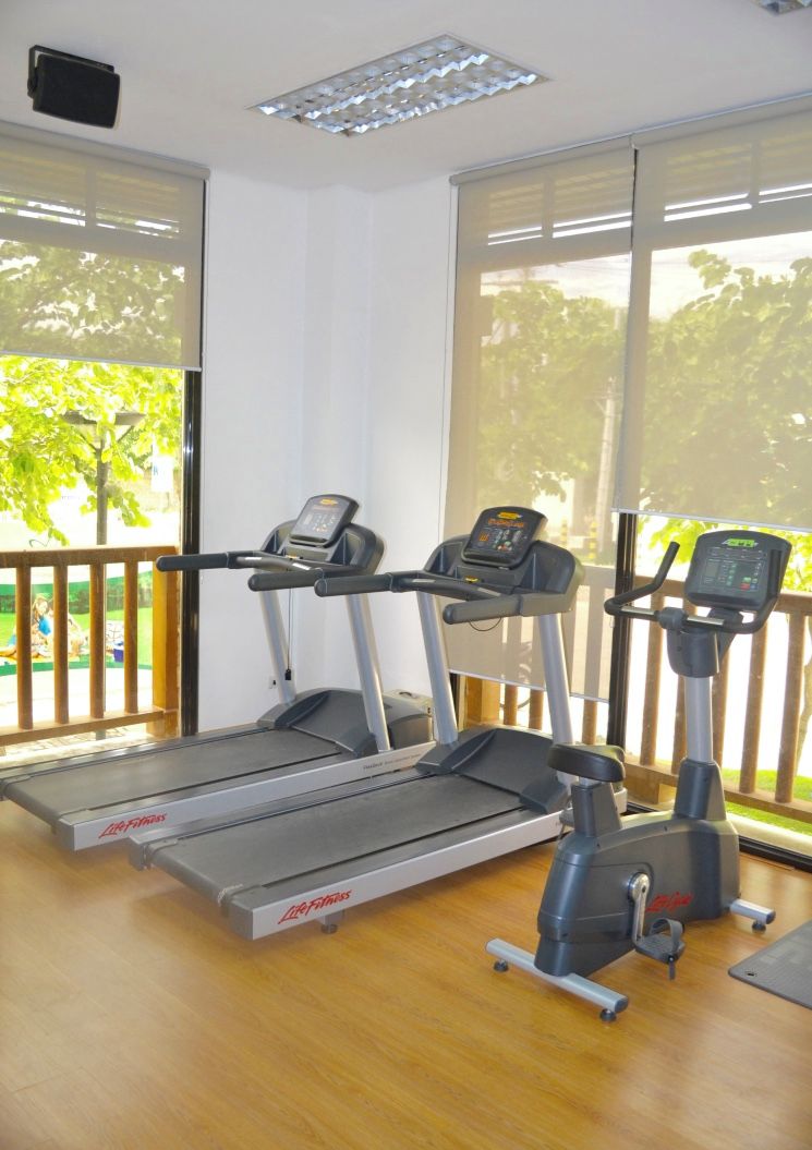 A gym with treadmills and an elliptical machine, large windows with a view of trees, and a wooden floor.