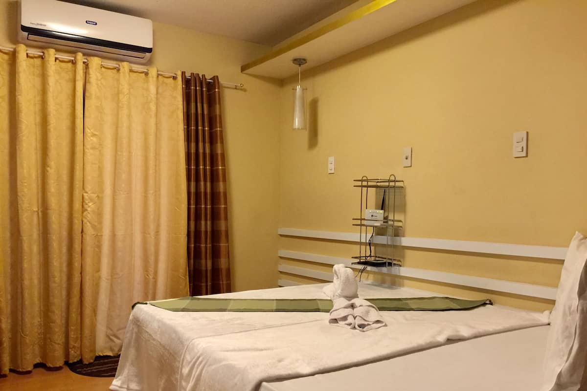 Une chambre d’hôtel comprenant un lit, une table de chevet, un appareil de climatisation, une étagère fixée au mur et des rideaux.