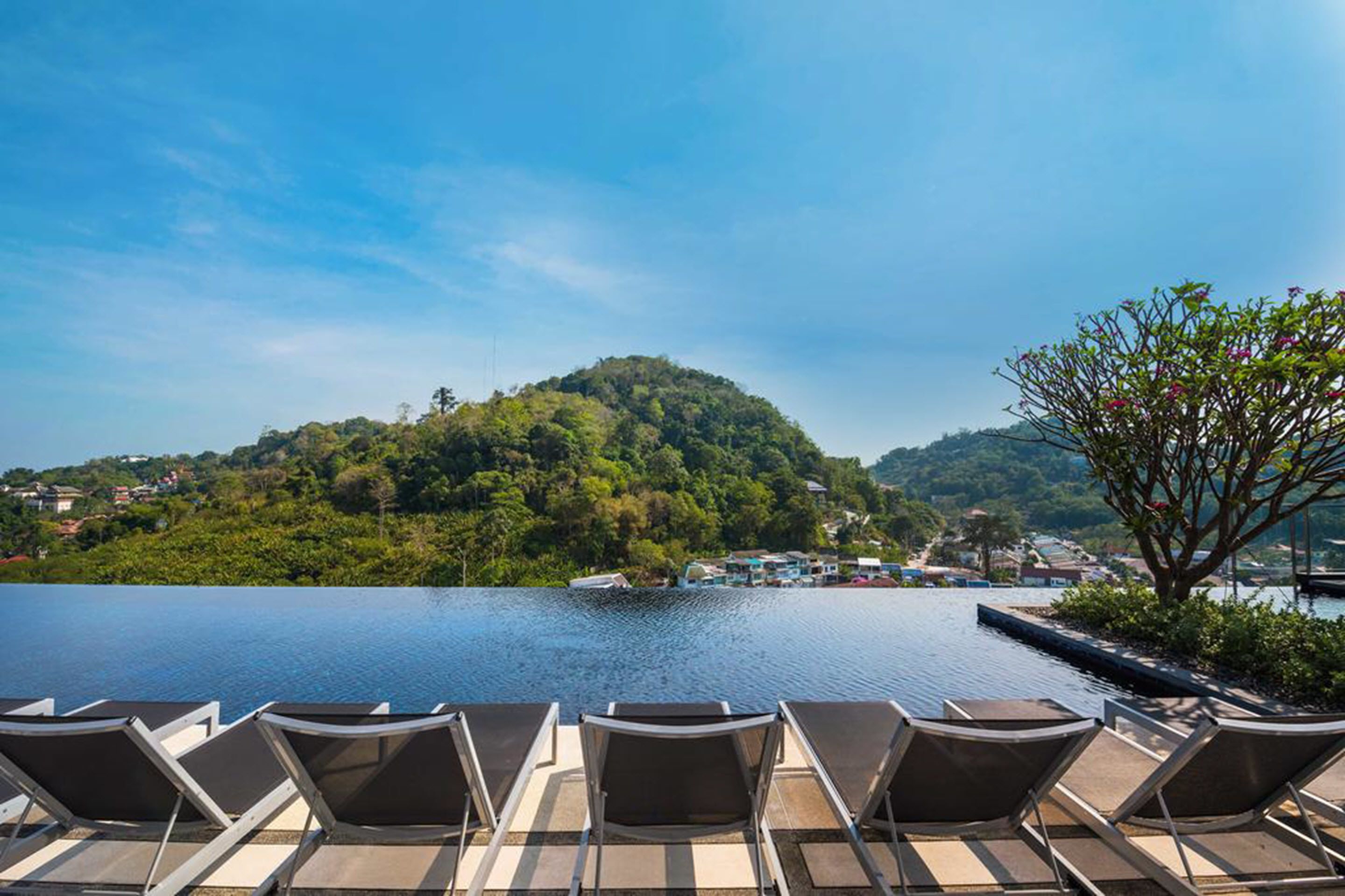 A poolside area with lounge chairs, a view of a lake, and a hill with greenery and buildings in the background.