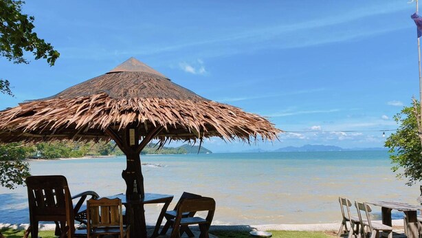 Un área de estar frente a la playa con una sombrilla de paja, sillas de madera y una mesa, con vista a un mar tranquilo y montañas lejanas.