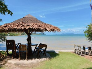 A beachside seating area with a thatched umbrella, wooden chairs, and a table, overlooking a calm sea and distant mountains. - Sabai Corner Bungalows (Ko Yao)