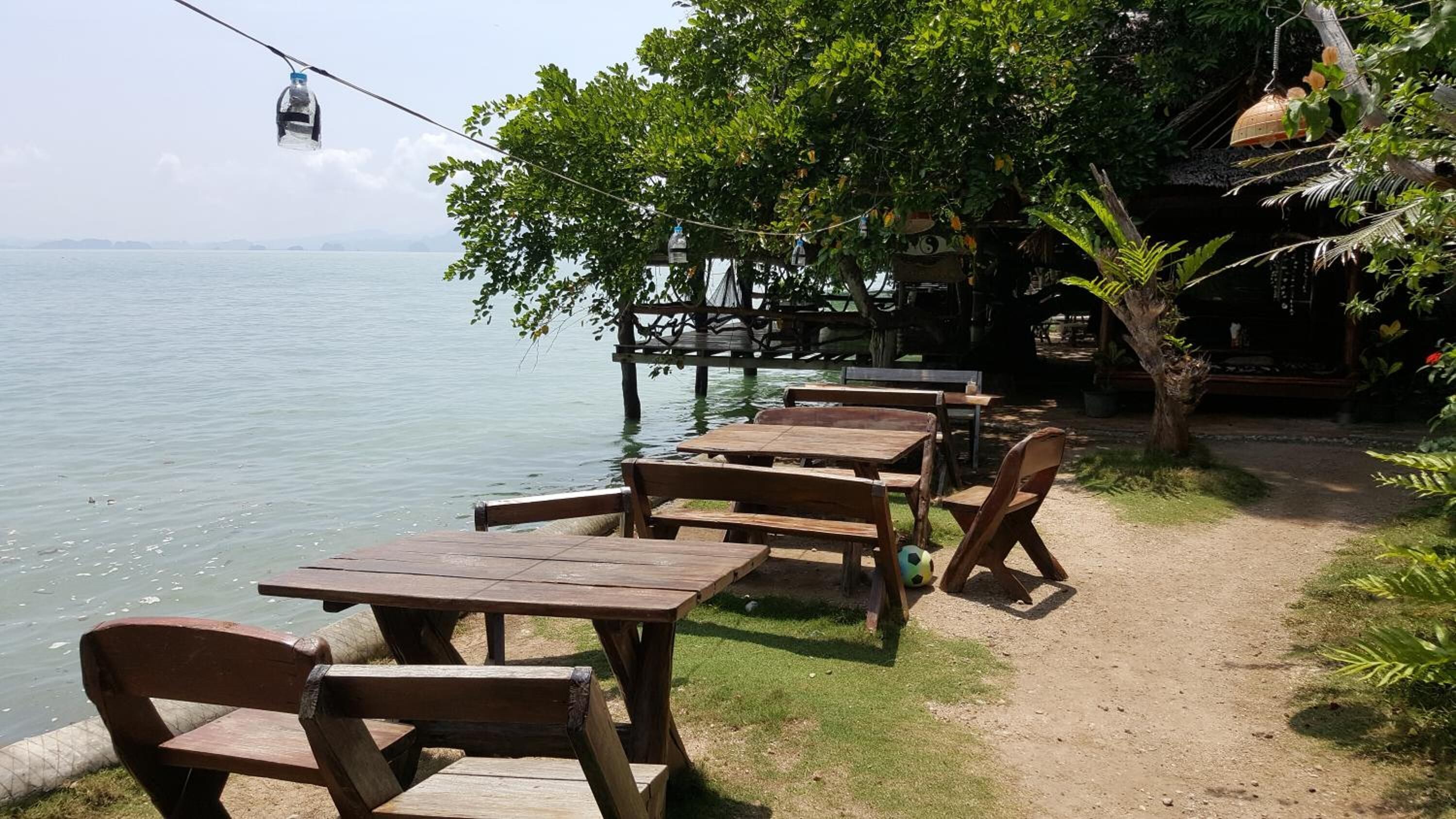 A seaside restaurant with wooden tables and benches, a view of the water, and a tree-lined path.