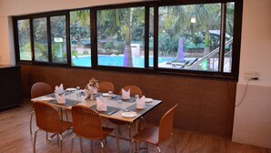 A dining area with a table set for four, large windows with a view of a swimming pool and tropical plants. - Clarissa Resort (Ramnagar)