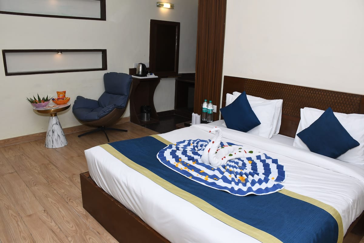 Cama bem arrumada com manta azul e branca, mesinha de cabeceira com uma planta e uma cadeira em um quarto de hotel.