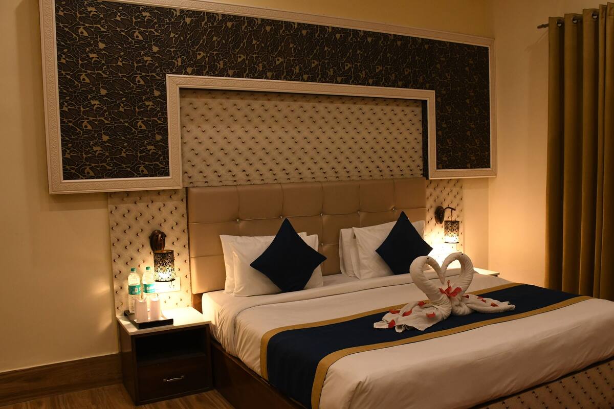 Quarto de hotel com cama grande, cabeceira com design estampado, mesa de cabeceira com abajur e cortina.