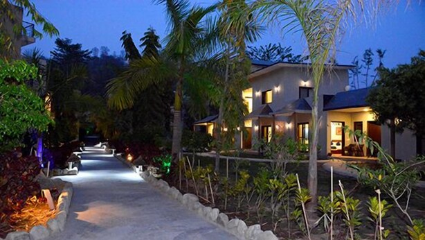 A resort with a paved pathway leading to a building with multiple rooms, surrounded by tropical vegetation and palm trees. - Clarissa Resort (Ramnagar)