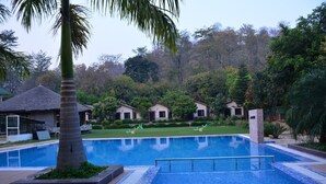 A swimming pool with a wooden deck, surrounded by lush greenery and small houses with thatched roofs. - Clarissa Resort (Ramnagar)