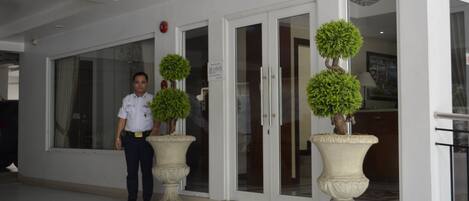 A hotel entrance with a receptionist, a glass door, and two large planters with topiary trees.